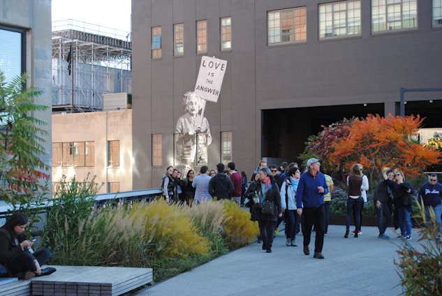 A group of people is gathered in an urban outdoor space featuring plants and trees with autumn foliage. A mural of a figure holding a sign that reads 'Love is the answer' is painted on the side of a building. The scene includes a mix of individuals either walking or standing, engaging in conversation, or using their phones, with a background of city architecture.