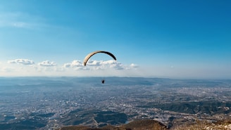 A paraglider with an orange and yellow wing is soaring above an expansive landscape that includes a detailed cityscape surrounded by hills and mountains. The sky is clear and blue with a few scattered clouds, adding a sense of freedom and openness to the scene.