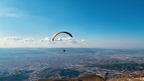 A paraglider with an orange and yellow wing is soaring above an expansive landscape that includes a detailed cityscape surrounded by hills and mountains. The sky is clear and blue with a few scattered clouds, adding a sense of freedom and openness to the scene.