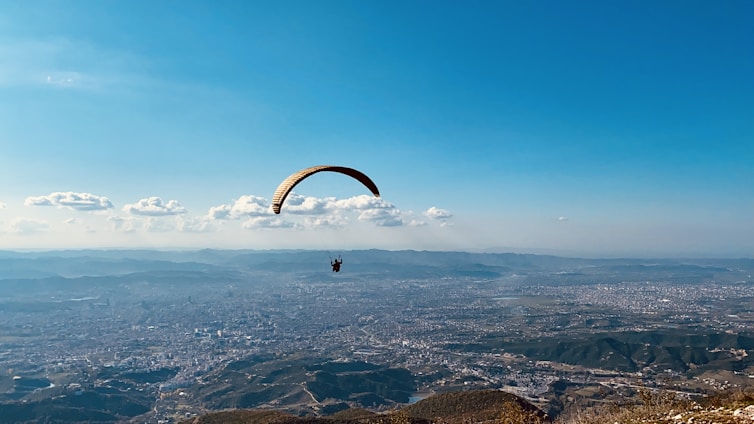 A paraglider with an orange and yellow wing is soaring above an expansive landscape that includes a detailed cityscape surrounded by hills and mountains. The sky is clear and blue with a few scattered clouds, adding a sense of freedom and openness to the scene.