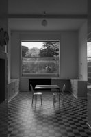 A minimalist kitchen features a checkered tile floor with a simple rectangular table and two chairs. Large windows frame a view of lush trees outside, providing natural light. The room is devoid of clutter, with cabinets lining the walls and a hanging light fixture in the center.