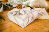 A neatly wrapped gift in a beige plaid fabric is topped with a small pinecone, placed on a wooden surface. In the background, there are blurred elements that suggest a natural or festive setting, possibly involving more gifts or decorative items.