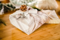 A neatly wrapped gift in a beige plaid fabric is topped with a small pinecone, placed on a wooden surface. In the background, there are blurred elements that suggest a natural or festive setting, possibly involving more gifts or decorative items.
