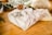 A neatly wrapped gift in a beige plaid fabric is topped with a small pinecone, placed on a wooden surface. In the background, there are blurred elements that suggest a natural or festive setting, possibly involving more gifts or decorative items.