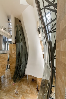 The interior of a modern architectural building with large, curving white and glass structures. The design features a blend of smooth walls and elongated windows, creating a futuristic and innovative atmosphere. People are walking and sitting around the spacious area on the warm-toned floor.