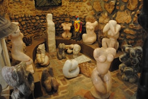 A variety of abstract stone sculptures, predominantly depicting stylized human torsos, are arranged in a circular manner within a rustic setting that features a stone wall. The sculptures vary in color and texture, with one brightly colored, patterned figure standing out among the more muted, natural stone hues.