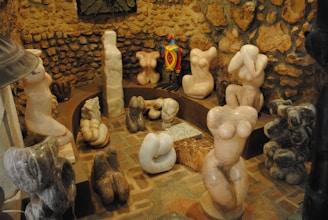 A variety of abstract stone sculptures, predominantly depicting stylized human torsos, are arranged in a circular manner within a rustic setting that features a stone wall. The sculptures vary in color and texture, with one brightly colored, patterned figure standing out among the more muted, natural stone hues.