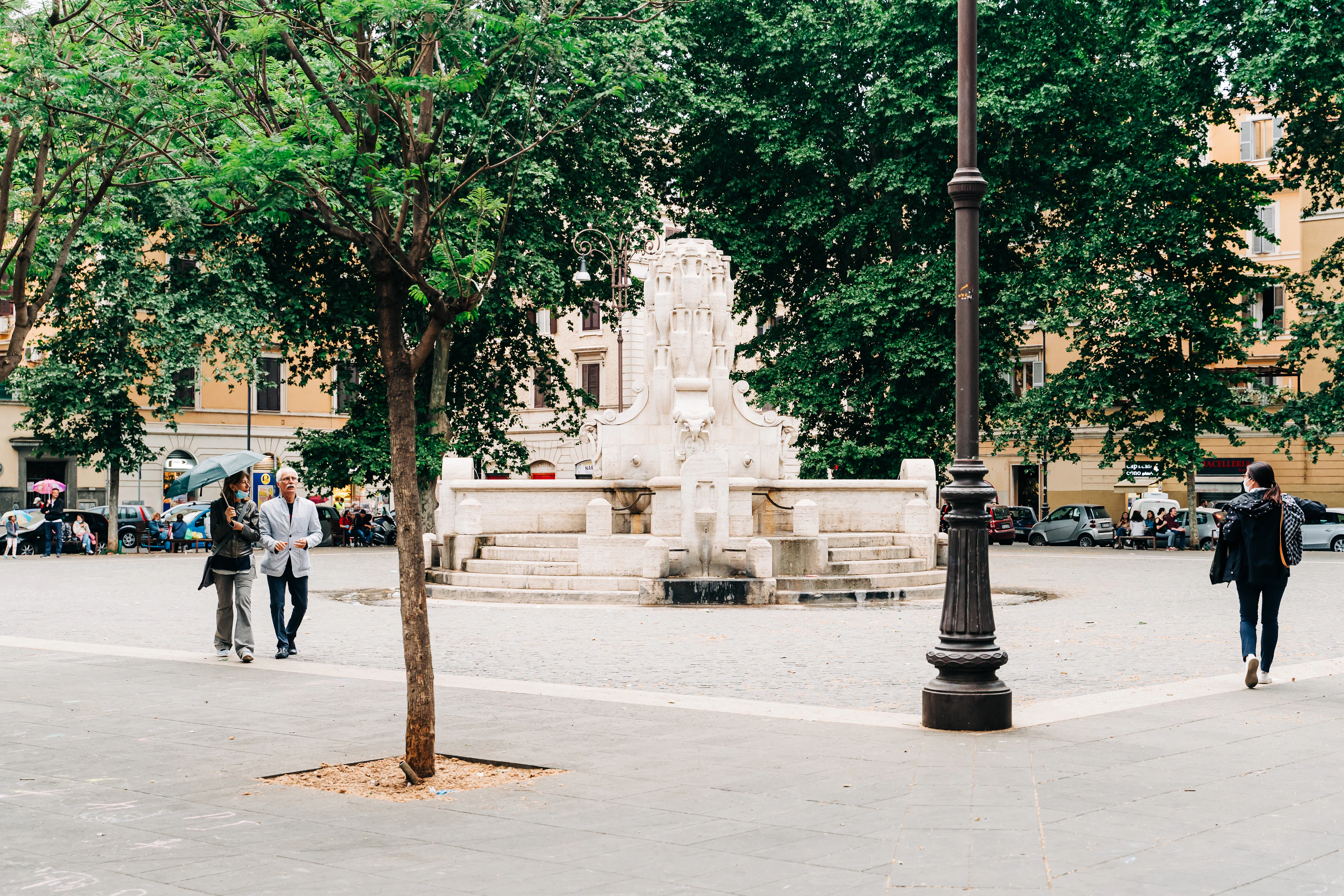 A small Roman piazza with a marble monument at its centre, plane trees and shuttered facades around it