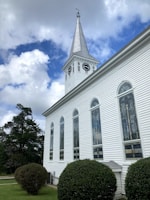 A white church building with tall, arched windows and a clock tower featuring two clocks on opposite sides. The tower has a pointed steeple with a weather vane on top. The church is surrounded by neatly trimmed hedges and trees, set against a backdrop of a partly cloudy sky with patches of blue.