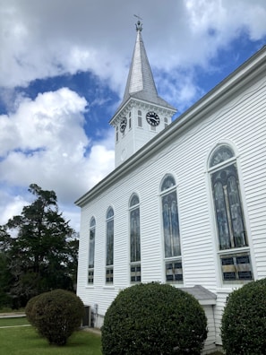 A white church building with tall, arched windows and a clock tower featuring two clocks on opposite sides. The tower has a pointed steeple with a weather vane on top. The church is surrounded by neatly trimmed hedges and trees, set against a backdrop of a partly cloudy sky with patches of blue.