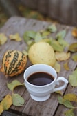 A steaming cup of coffee resting on a wooden table surrounded by pinecones and autumn leaves.