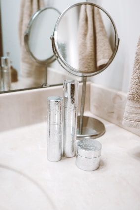 A clean bathroom countertop features silver cosmetic containers placed neatly next to a round tabletop mirror. The mirror has a stand and is complemented by beige towels hanging nearby. The setting appears organized and minimalist.