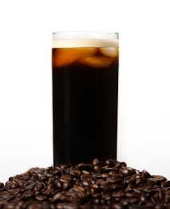 A tall glass filled with a dark beverage topped with a layer of foam and ice cubes is surrounded by a pile of glossy coffee beans. The background is plain and light, providing contrast to the dark tones of the drink and beans.