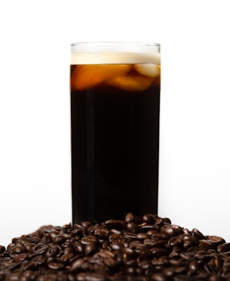 A tall glass filled with a dark beverage topped with a layer of foam and ice cubes is surrounded by a pile of glossy coffee beans. The background is plain and light, providing contrast to the dark tones of the drink and beans.