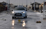 A white car with a driving school sign on top is driving through a wet street. Debris and puddles are scattered across the road. People are walking on the sidewalk, wearing jackets, suggesting cold or rainy weather. Benches and street lamps line the road, with a gazebo-like structure visible in the background.