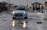 A white car with a driving school sign on top is driving through a wet street. Debris and puddles are scattered across the road. People are walking on the sidewalk, wearing jackets, suggesting cold or rainy weather. Benches and street lamps line the road, with a gazebo-like structure visible in the background.