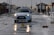 A white car with a driving school sign on top is driving through a wet street. Debris and puddles are scattered across the road. People are walking on the sidewalk, wearing jackets, suggesting cold or rainy weather. Benches and street lamps line the road, with a gazebo-like structure visible in the background.