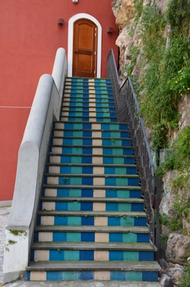 A staircase with alternating blue and beige tiles leads up to a wooden door set in a red wall. The right side of the staircase is bordered by black wrought iron railing, while the left side features a smooth, white concrete banister. Greenery grows alongside the rock face to the right of the stairs.
