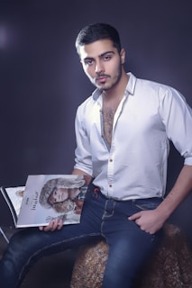 A man with short dark hair and facial hair is wearing a white button-up shirt with the top buttons undone, paired with dark blue jeans. He is sitting on a textured stool, holding an open magazine featuring a model with a fur hood.
