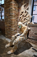 A mannequin made from paper sits on a chair, holding a blue book. It is surrounded by a large stack of books and wall decorations crafted from open books, arranged to create a sculptural effect. The setting is a room with exposed brick walls and a window, contributing to a rustic and artistic atmosphere.