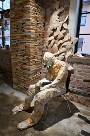 A mannequin made from paper sits on a chair, holding a blue book. It is surrounded by a large stack of books and wall decorations crafted from open books, arranged to create a sculptural effect. The setting is a room with exposed brick walls and a window, contributing to a rustic and artistic atmosphere.