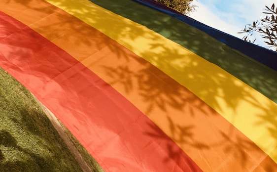 A vibrant, multi-colored fabric with horizontal stripes of red, orange, yellow, green, blue, and purple, laid out on a grassy area. Sunlight casts leaf shadows on the fabric, adding a natural pattern to the composition. A portion of the sky and tree branches are visible at the top edge.