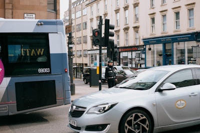 A city street scene featuring a bus and a car stopped at a traffic light. A traffic officer stands on the corner, monitoring the intersection. Surrounding buildings have various businesses, including a beauty salon and an acupuncture clinic.
