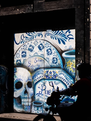 A vibrant mural featuring a large, ornate skull painted with intricate blue and white patterns, inspired by traditional Mexican tile and Day of the Dead motifs. The artwork includes floral designs and the words 'Se&ntilde;or Housing' and 'Puebla' incorporated into the design. The scene is partly obscured by shadows and a silhouette of a person on a motorcycle in the foreground.