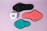 Three reusable sanitary pads in different colors are laid out on a pastel purple background. The colors include black, coral pink, and teal, each with black borders. A box with the branding 'inciclo' is placed beside the pads, featuring a design with teal shapes and floral elements.