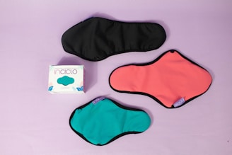 Three reusable sanitary pads in different colors are laid out on a pastel purple background. The colors include black, coral pink, and teal, each with black borders. A box with the branding 'inciclo' is placed beside the pads, featuring a design with teal shapes and floral elements.