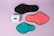 Three reusable sanitary pads in different colors are laid out on a pastel purple background. The colors include black, coral pink, and teal, each with black borders. A box with the branding 'inciclo' is placed beside the pads, featuring a design with teal shapes and floral elements.