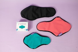 Three reusable sanitary pads in different colors are laid out on a pastel purple background. The colors include black, coral pink, and teal, each with black borders. A box with the branding 'inciclo' is placed beside the pads, featuring a design with teal shapes and floral elements.
