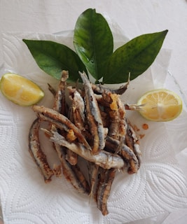A pile of fried small fish arranged on a white paper towel, accompanied by two lemon wedges and large fresh green leaves for garnish.