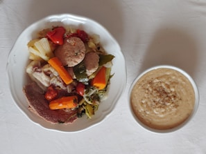 A plate filled with a variety of cooked vegetables and meats, including carrots, red peppers, and possibly chicken or beef, accompanied by a bowl of creamy soup or sauce with pepper sprinkled on top. The setting is simple with a light-colored tablecloth.
