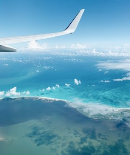 An aerial view of an ocean landscape featuring a mix of turquoise and deep blue waters. A white airplane wing is visible on the left side, partially in the frame. Fluffy white clouds are scattered across the sky, and a thin strip of land with sandy beaches is in the distance.