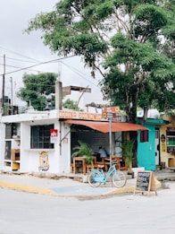 A small, rustic restaurant with a sign that reads 'Cocina Económica'. A teal bicycle is parked in front, and there are wooden tables and chairs arranged outside under a canopy. The establishment is adjacent to a large tree and there are handwritten menu items displayed on a small chalkboard.
