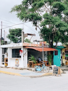 A small, rustic restaurant with a sign that reads 'Cocina Económica'. A teal bicycle is parked in front, and there are wooden tables and chairs arranged outside under a canopy. The establishment is adjacent to a large tree and there are handwritten menu items displayed on a small chalkboard.