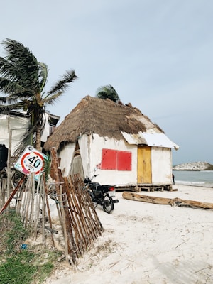 A small beach hut with a thatched roof stands on a sandy shore, surrounded by palm trees swaying in the wind. A motorcycle is parked beside the hut, and a weathered traffic sign indicating a speed limit of 40 km/h is leaning against a wooden fence. The sky is overcast, and the ocean is visible in the background.