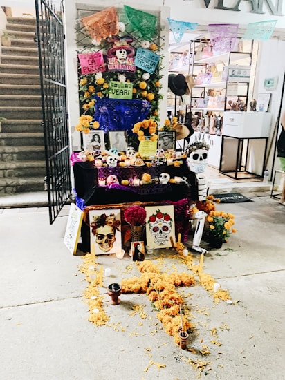 An ornate altar decorated for D&iacute;a de los Muertos with marigold flowers, colorful papel picado banners, and an arrangement of candles and skulls. Photographs of deceased loved ones are displayed, along with painted skull artwork and other offerings.