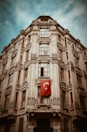 A historic multi-story building featuring ornate architectural details and large windows, with a Turkish flag hanging prominently on the facade. The sky is a clear blue, creating a striking contrast with the building's stone exterior.