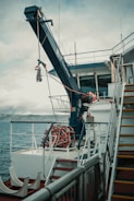 A ship deck with a large crane labeled 'Global Davit' reaching over the side. A neatly coiled rope or hose is visible on the deck. The railing, stairs, and some equipment are also present. The background shows the sea and a distant mountain range under a cloudy sky.