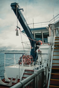 A ship deck with a large crane labeled 'Global Davit' reaching over the side. A neatly coiled rope or hose is visible on the deck. The railing, stairs, and some equipment are also present. The background shows the sea and a distant mountain range under a cloudy sky.