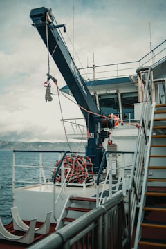 A ship deck with a large crane labeled 'Global Davit' reaching over the side. A neatly coiled rope or hose is visible on the deck. The railing, stairs, and some equipment are also present. The background shows the sea and a distant mountain range under a cloudy sky.
