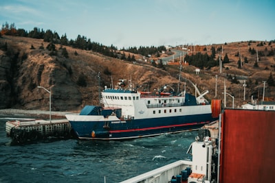 A ferry is docked by a rugged coastline with hills covered in sparse vegetation. The water is a deep blue-green, and there is a small pier with mooring structures. In the background, paved roads wind up the hillside, accompanied by utility poles and a few scattered buildings.