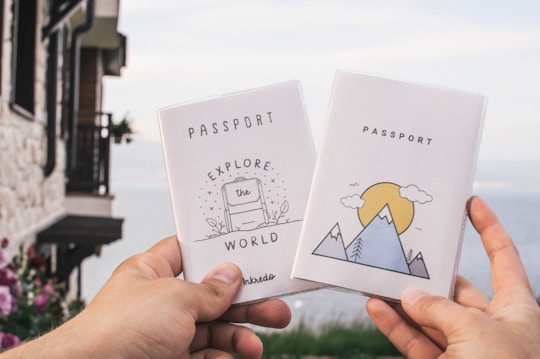 Two hands are holding up passport covers against the backdrop of a scenic lake. The left cover has an illustration of a suitcase with the words 'EXPLORE the WORLD'. The right cover features a mountain and sun illustration. There is a stone building with a rustic look on the left and flowering plants in the bottom left.
