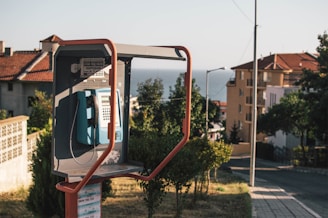 An outdoor telephone booth located on a street with houses in the background. The booth contains a blue public phone, and a mix of modern and traditional architecture can be seen. Vegetation and trees are present, and the view extends to a distant sea under a clear sky.