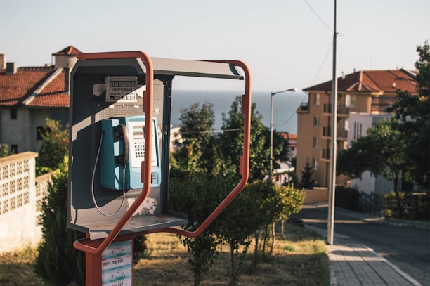 An outdoor telephone booth located on a street with houses in the background. The booth contains a blue public phone, and a mix of modern and traditional architecture can be seen. Vegetation and trees are present, and the view extends to a distant sea under a clear sky.