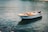 A small wooden boat is floating on calm water, tethered by ropes at the front. The boat has a traditional design with a painted exterior and an engine attached at the rear. The surrounding water is a clear blue-green, and a few buoys can be seen in the distance. The background includes some rocky edges.