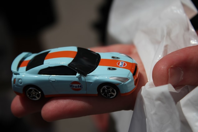 A small toy car with a Gulf oil company logo held in a person's hand. The car features a light blue body with an orange stripe running across its length. It is being held gently, with part of a white tissue or cloth visible underneath the hand.