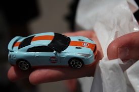 A small toy car with a Gulf oil company logo held in a person's hand. The car features a light blue body with an orange stripe running across its length. It is being held gently, with part of a white tissue or cloth visible underneath the hand.